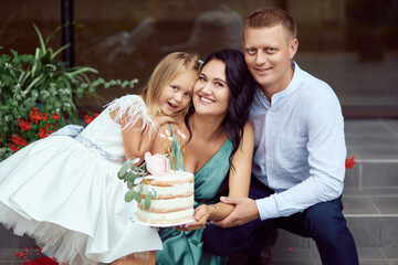 Parents with their daughter celebrating a birthday on the street, a cake in their hands. Happy family together.