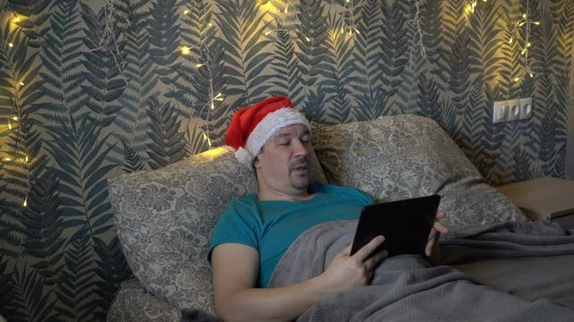 A sick adult male communicates via video link via a tablet while lying at home on a bed in a Santa Claus hat. The concept of celebrating Christmas during the coronavirus pandemic. 4K