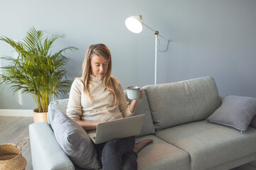 Young woman enjoying her coffee while working or studying on laptop computer at home office. Smiling young holding a cup of coffee while working on her laptop in her home office