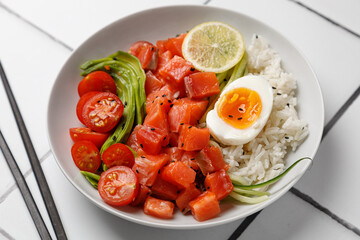 Salmon Poke bowl with avocado, cherry, egg and rice on white tile background with chopsticks. Top view
