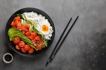 Top view of poke bowl with salmon, avocado and rice on dark background with chopsticks