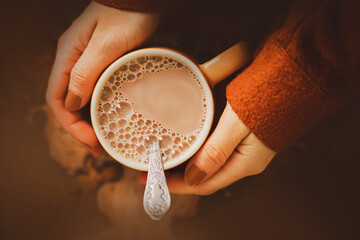 A woman in a brown woolen jacket is holding a mug of warm fragrant delicious cocoa with milk. A...