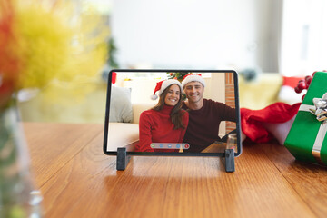 Happy caucasian couple in santa hats smiling on tablet video call screen at christmas time