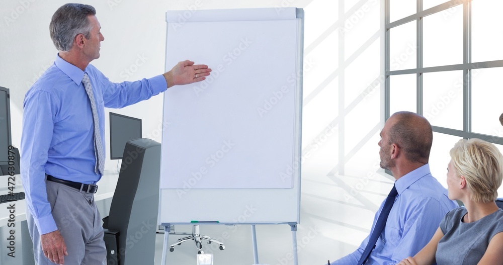 Wall mural Mature businessman gesturing at blank flipchart with copy space to colleagues during meeting