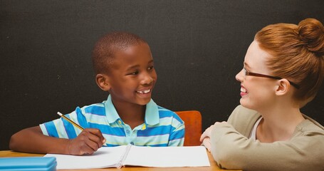Smiling schoolboy with book and pencil looking at happy redhead teacher in classroom