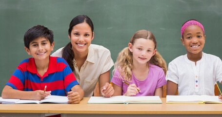Portrait of smiling teacher with schoolboy and schoolgirls at desk against blackboard in classroom