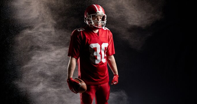 Male american football player in red uniform holding ball against black background with mist