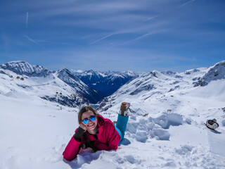 Fototapeta na wymiar A girl in a snowboarding outfit lies on the snow and enjoys her break. Endless chains of snow caped Alps surrounding the girl. Girl is smiling. Clear and bright day. Moelltaler Gletscher, Austria.