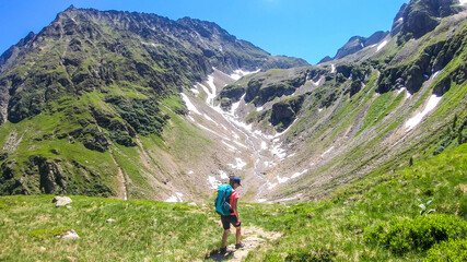 A young woman with a big backpack hikes down on a steep pathway between tall mountain peaks. Some of the slopes are covered with snow. In the back is another mountain range. Spring in alpine valleys.