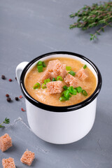 Vegetable cream soup topped with croutons and scallion served in the metallic mug on the gray table. Hot meal