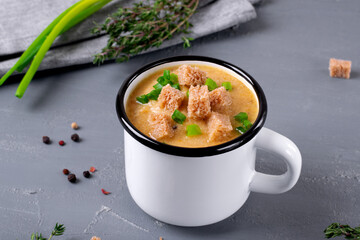 Vegetable cream soup topped with croutons and scallion served in the metallic mug on the gray table. Hot meal