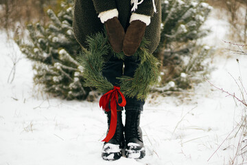 Stylish woman with christmas wreath in winter park. Young female in cozy mittens and coat holding...