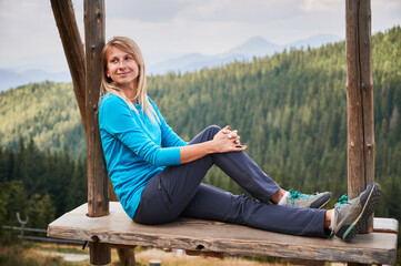 Nice girl resting on wooden seat suspended on two wooden poles and enjoying amazing scenery. On the background green forest and silhouettes of mountain hills far away.