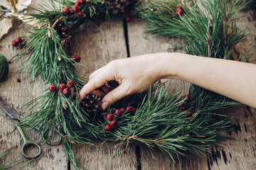 Woman making Christmas wreath by attaching red berries to fir tree branch during Christmas...