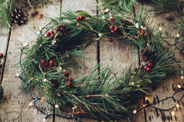Handmade Christmas wreath near mistletoe on wood table closeup, Christmas lights around festive wreath with pinecones and berries, scandinavian home decor