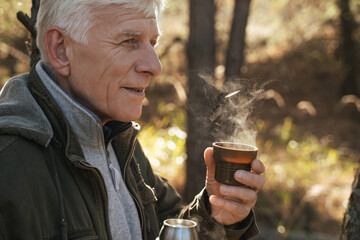 Positive tourist holding cup with hot tea while sitting on the ground in pinewood