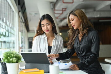 Photo of young businesswomen working together at the modern counter bar surrounded by a corporate document, digital tablet and office equipment.