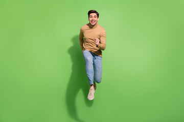 Photo of energetic sportive guy run forward jump wear beige shirt jeans sneakers isolated green...