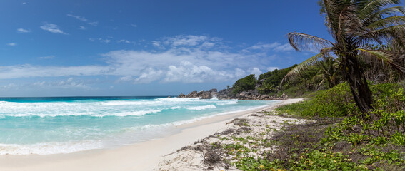 A high-resolution panorama of a coastal landscape with wide white sand beach in the foreground. Foamed ocean surf washes the shore. There are stoneformations and rainforest along the coastline.