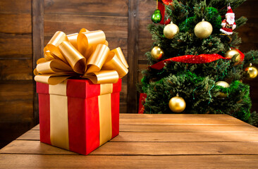 Christmas and New Year Red Box Gift Decorated by Golden Ribbon and Big Golden Loopy Bow. Christmas Tree with Balls, Ribbons, Santa Toy ,Serpentine and Garlands, Close-up. Wood Background