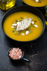 Bowl of pumpkin soup topped with soft cheese and pumpkin seeds, vertical shot on a dark-brown stone surface, middle close-up
