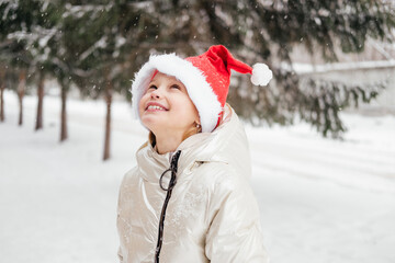 Smiling little girl in red christmas hat standing under snowfall looking up. Outdoors winter activities. Happy child in Santa hat playing, having fun in winter forest.