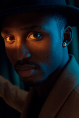 Portrait photo of a guy with dark skin. He has a black hat on his head. Photo taken on a dark background. The sun shines on his face and the model looks straight into the camera