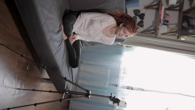 A woman blogger with red dreadlocks recording video or live broadcast using smartphone and tripod at home.Online show On-air production host streaming live content, recording digital social media