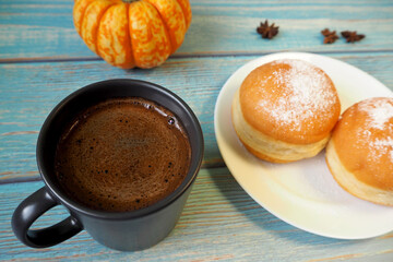 two doughnuts sprinkled with powdered sugar on a white plate next to a dark coffee mug and an orange pumpkin on a blue wooden background. side view. breakfast