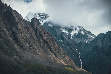 Fototapeta premium Awesome mountain landscape with snowy mountains in low clouds. Rocky top and snowy top. Atmospheric view to great rocky mountains in cloudy sky. Gloomy scenery with high mountains in overcast weather.