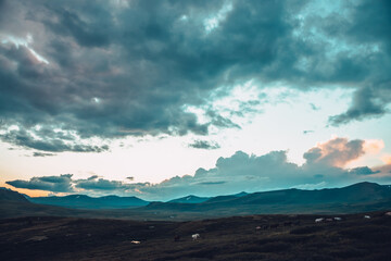 Surrealist mountain scenery with horses and golden dawn light in cloudy sky. Scenic mountain landscape with horses on hill and surreal sunset sky. Orange sunlight in sunrise sky. Fantasy mountain view