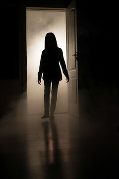 A silhouette of a little girl emerges from a luminous room with smoke in the dark.