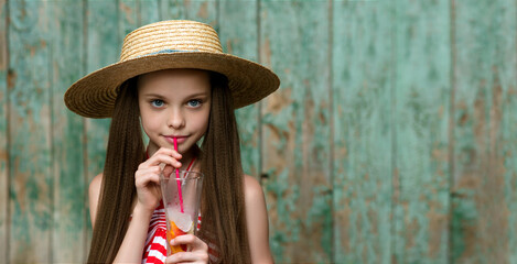 Cute little girl drinks lemonade front of old rural wooden background. Kid holds glass with summer...