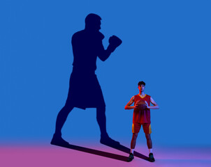 Fototapeta premium Young man dreaming about famous sport future. Conceptual image with sportive boy and shadow of boxer on blue wall