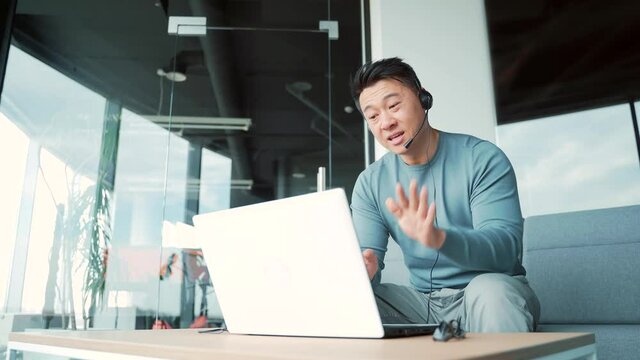 Asian male freelancer talking to colleagues in meeting room at modern office coworking. Business man conference online using laptop headset and webcam. man entrepreneur or an employee work remotely