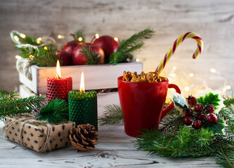 Red apples in a wooden box with fir branches and garlands. A red cup with cookies and lollipops. Red and green candles on a light wooden table. Festive atmosphere