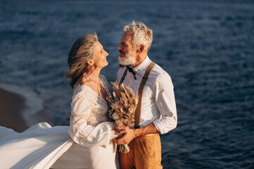 Senior newlyweds hug. Stylish couple of elderly newlyweds stand embracing on river bank.