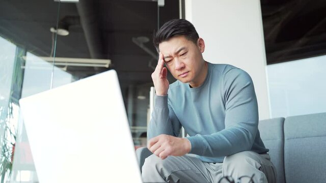 Young exhausted, overworked office worker sitting in the workplace with a severe headache massaging his head. Portrait unhealthy Tired Asian male in pain sick Stress at work sedentary working computer