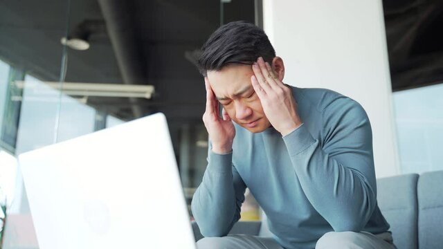 Young exhausted, overworked office worker sitting in the workplace with a severe headache massaging his head. Portrait unhealthy Tired Asian male in pain sick Stress at work sedentary working computer