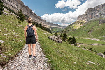 A woman walks through a mountain valley wearing summer clothes. The path ascends towards the top of the mountain.