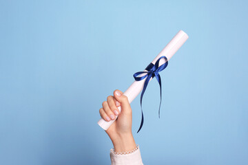 Student holding rolled diploma with ribbon on light blue background, closeup