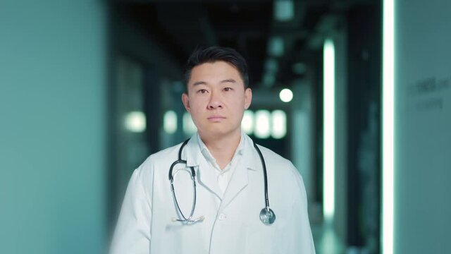 Asian male doctor walks down the hallway of a modern hospital clinic. Portrait of a serious confident medic therapist or surgeon going indoors hall