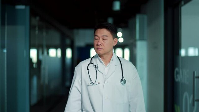 Asian male doctor walks down the hallway of a modern hospital clinic. Portrait of a serious confident medic therapist or surgeon going indoors hall