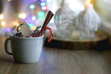 Mug filled with candy cane, cinnamon sticks and various Christmas decorations. Colorful bokeh lights in the background. Selective focus.