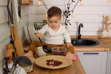 A little brunet boy, sits on the kitchen table, plays with cutlery and beans,