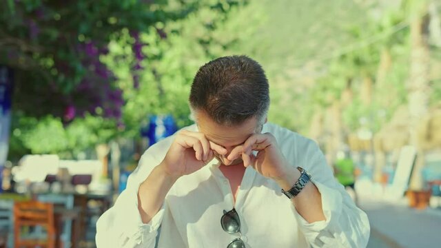 Young bearded man coughing while walking outside, feeling sick.Man with pollen allergy running tears, eyes itching and sneezing on a beautiful flowery road. The concept of living with allergies.