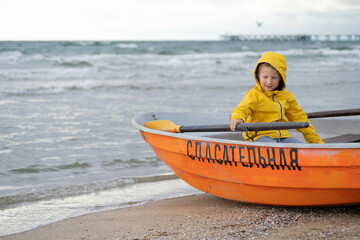When I grow up I will be a lifeguard. Boy in a yellow jacket sits in a lifeboat on the seashore. He has an oar in his hands. Cold winter day, cloudy weather, empty beach.