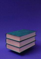 Three books, hardcover, stacked horizontally, one on top of the other, on a blue-violet background. Text space. Reading, leisure concept. Minimal style.