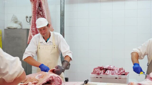 Front motion of male butcher sharpening knife at table and looking at camera. Concentrated man wearing uniform cutting skin off big meat piece at factory. Food industry concept.