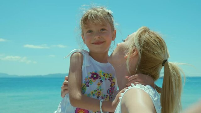 Blonde-haired mother kissing her little daughter, enjoying the holiday. Happy mother daughter concept.Front camera view.Little blonde-haired girl and her mother are taking a selfie on the beach.
